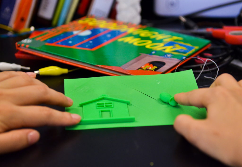 An image of a tactile graphic of a house and a mouse, 3D printed with green filament. Two hands touching the page.